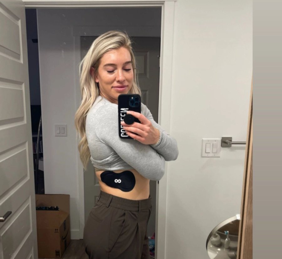 Woman in a grey long-sleeve top taking a mirror selfie, wearing an Ion Layer patch on her abdomen.