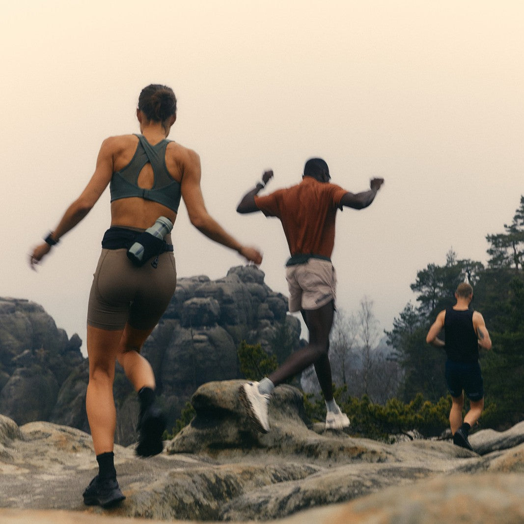 Three runners climbing a rocky ridge at dawn, with pine forest and low mist behind them.